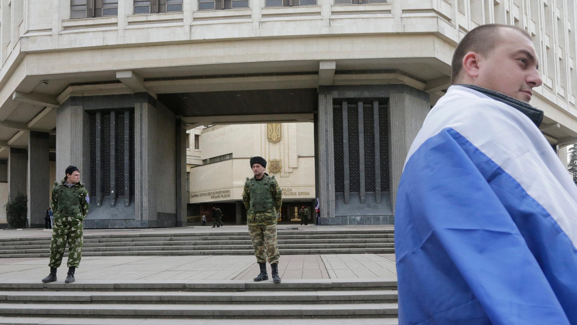 Ein in eine russische Flagge gehüllter prorussischer Aktivist am 6. Maärz 2014 vor dem Parlament der Krim, das von zwei prorussischen Uniformierten, vermutlich Kosaken bewacht wird.