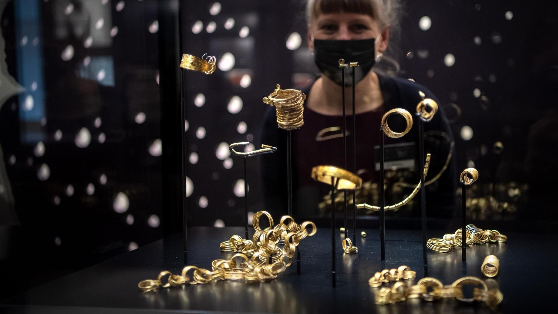 Eine Frau mit Mund-Nasen-Schutz steht im Kreismuseum Syke hinter einer Vitrine, in welcher der Goldhort von Gessel ausgestellt ist (gestellte Szene).