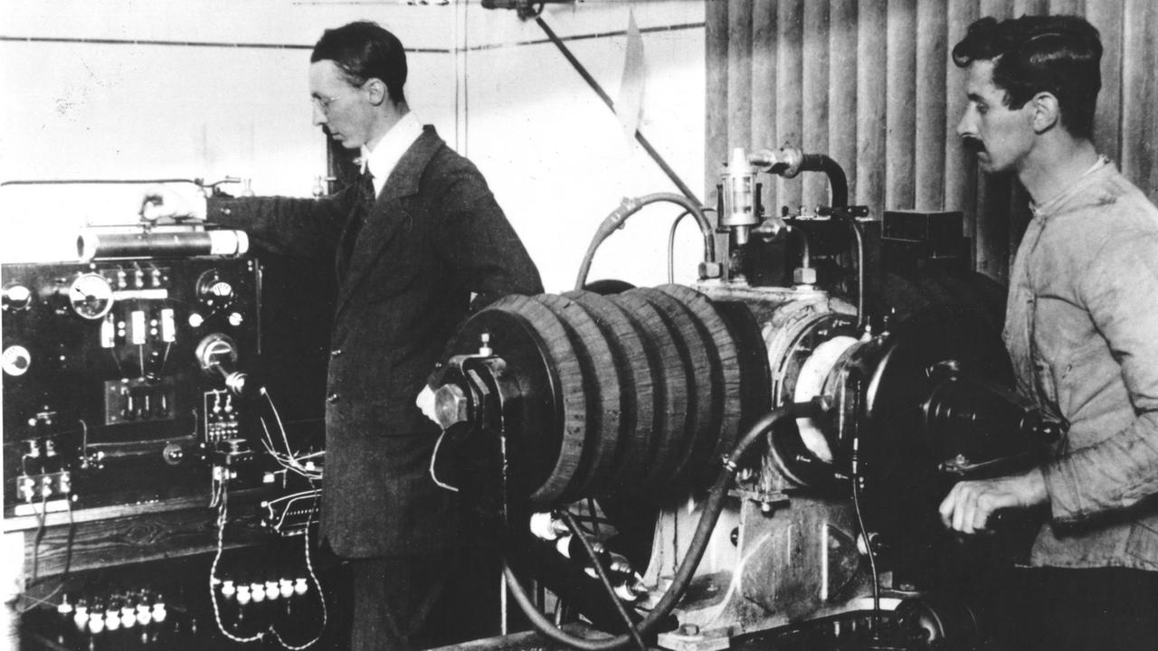 Auf einer historischen Fotografie sind zwei Männer zu sehen, die an technischen Geräten beschäftigt sind. Auf einer historischen Fotografie sind zwei Männer zu sehen, die an technischen Geräten beschäftigt sind.