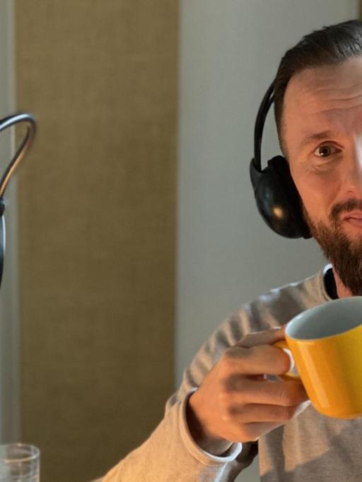 Robert Marc Lehmann am Lesepult vor dem Studio-Mikrofon, mit Kopfhörern auf den Ohren und einer gelben Kaffeetasse in der Hand