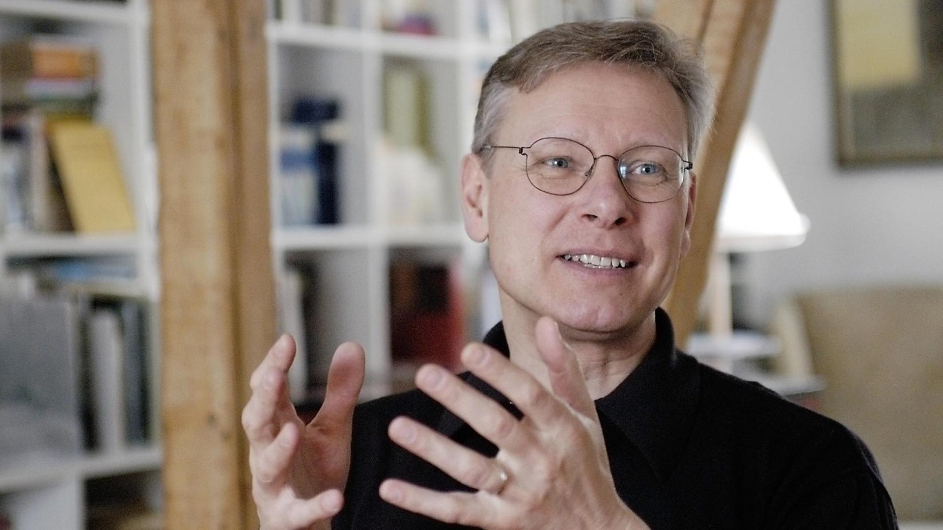 Ein mittelalter Mann mit Brille gestikuliert beim Reden mit den Händen. Im Hintergrund ist ein Bücherschrank zu sehen. Ein mittelalter Mann mit Brille gestikuliert beim Reden mit den Händen. Im Hintergrund ist ein Bücherschrank zu sehen.