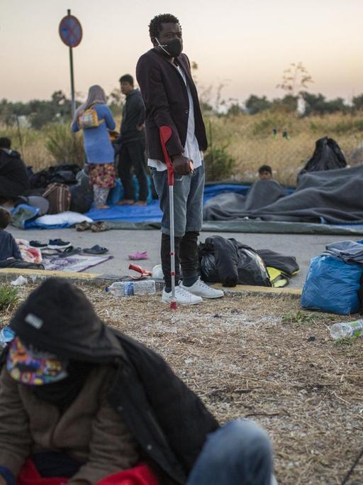 Menschen liegen und stehen am Rande einer Straße in der Nähe des ausgebrannten Flüchtlingslagers Moria aus.