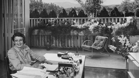 Die Schriftstellerin Ayn Rand sitzt an ihrem Schreibtisch in ihrem von Richard J. Neutra entworfenen Haus im San-Fernando-Tal; sie lächelt und schaut mit den Augen von der Kamera weg; sie trägt eine grau geknöpfte Bluse und hat beide Hände auf dem Schreibtisch; hinter ihr befindet sich ein Deck mit Kastenpflanzen und ein Metallstuhl mit einem Kissen und dem Blick auf das Tal.