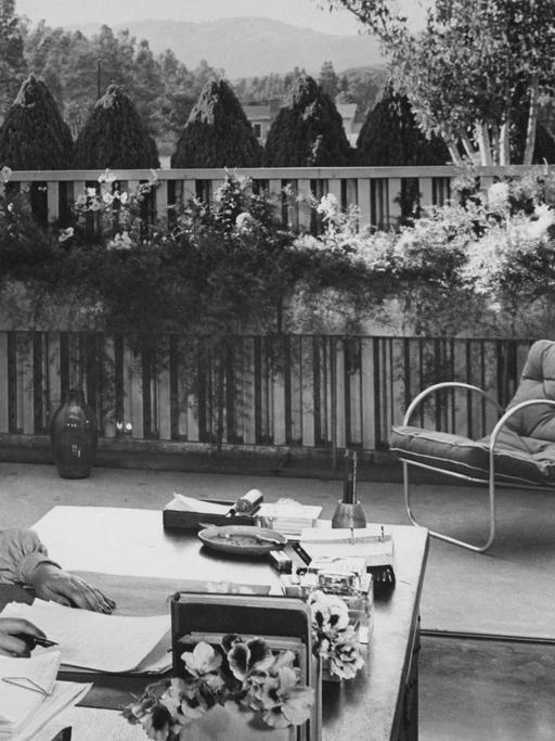 Die Schriftstellerin Ayn Rand sitzt an ihrem Schreibtisch in ihrem von Richard J. Neutra entworfenen Haus im San-Fernando-Tal; sie lächelt und schaut mit den Augen von der Kamera weg; sie trägt eine grau geknöpfte Bluse und hat beide Hände auf dem Schreibtisch; hinter ihr befindet sich ein Deck mit Kastenpflanzen und ein Metallstuhl mit einem Kissen und dem Blick auf das Tal.
