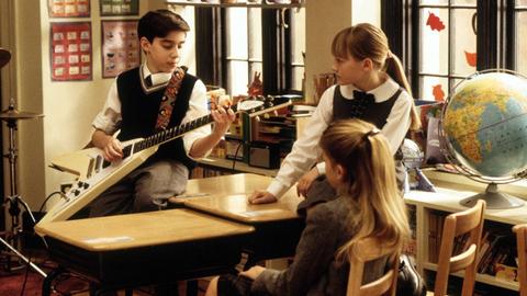Szene aus der Komödie "School of Rock": Ein Junge in Schuluniform spielt vor zwei Mitschülerinnen im Klassenraum auf einer E-Gitarre.