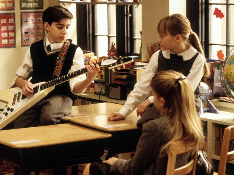 Szene aus der Komödie "School of Rock": Ein Junge in Schuluniform spielt vor zwei Mitschülerinnen im Klassenraum auf einer E-Gitarre.