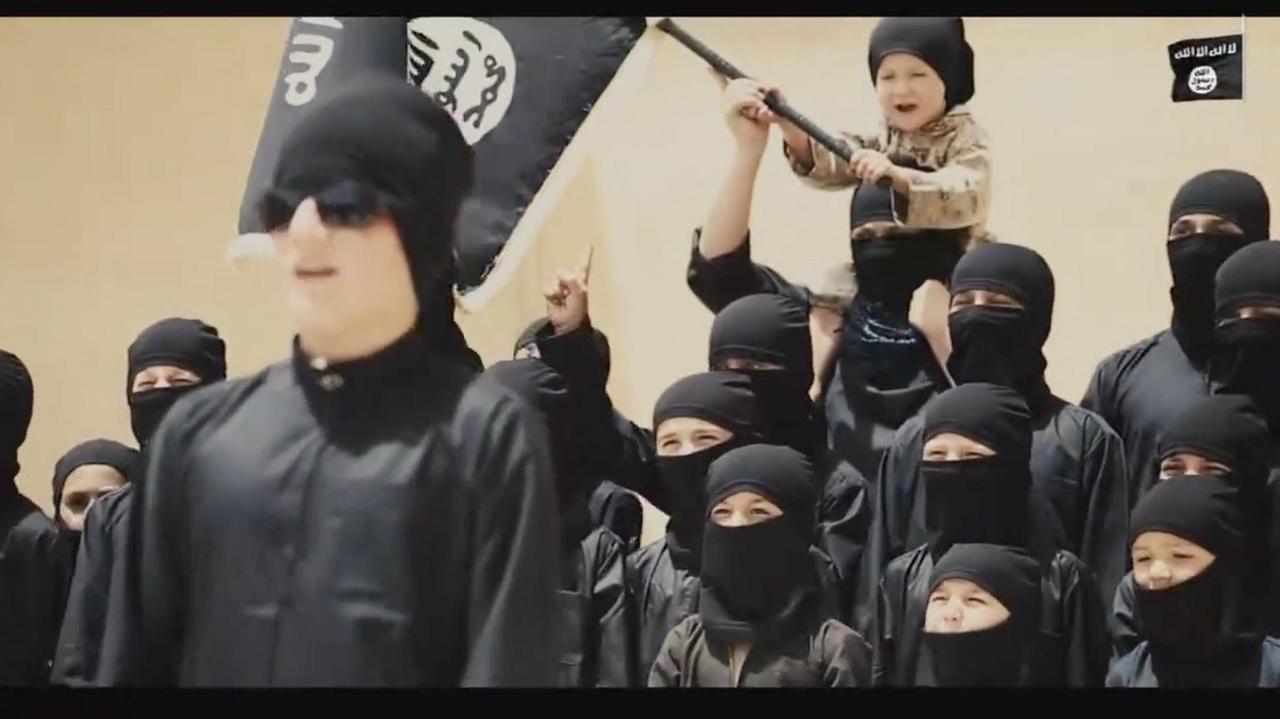 Das Bild zeigt schwarz vermummte Kinder, eines davon hält eine IS-Fahne. Vor ihnen steht ein erwachsener IS-Mann.