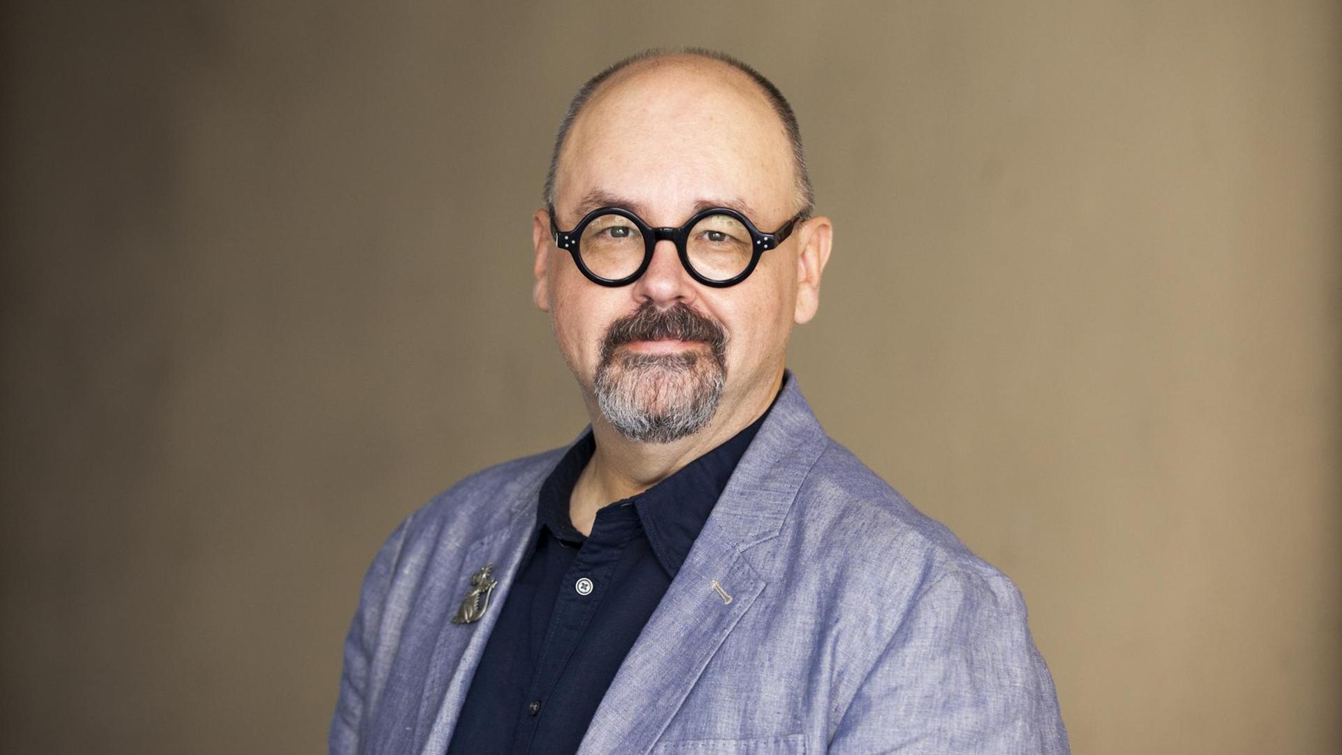 Carlos Ruiz Zafón trägt eine schwarze Hornbrille, dunkelblaues Polo und hellblaues Leinensakko vor gelblichem Hintergrund. Er schaut mit einem kleinen Lächeln direkt in die Kamera. Carlos Ruiz Zafón trägt eine schwarze Hornbrille, dunkelblaues Polo und hellblaues Leinensakko vor gelblichem Hintergrund. Er schaut mit einem kleinen Lächeln direkt in die Kamera.