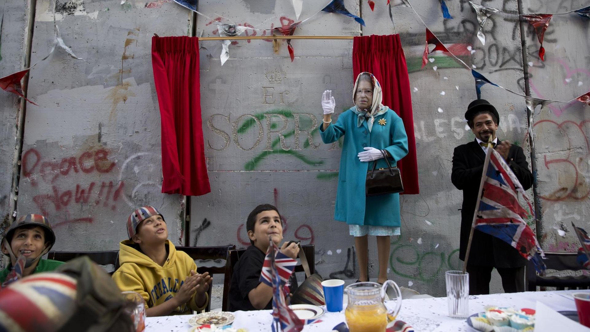 Kinder und eine als Königin Elizabeth verkleidete Person bei einem Event des britischen Künstlers Banksy.