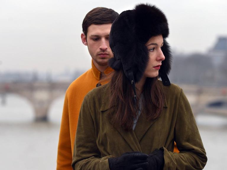 Eine Frau mit Pelzmütze steht vor einem Mann mit einer orange-fabenen Jacke, hinter ihnen die Seine in Paris