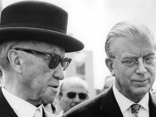 Kanzler Konrad Adenauer trägt eine Sonnenbrille und einen schwarzen Hut, rechts neben ihm steht Kanzleramtschef Hans Globke