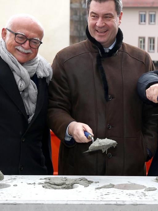 Gerd Schmelzer (l-r), Bauunternehmer aus Nürnberg, Markus Söder (CSU), Ministerpräsident von Bayern und Wolfgang Heckl, Generaldirektor des Deutschen Museums, stehen auf dem Gelände des früheren Augustinerhofs nebeneinander. Jeder von ihnen hat eine Kelle mit Speis darauf in der Hand. Sie schauen freudig in die Kamera.