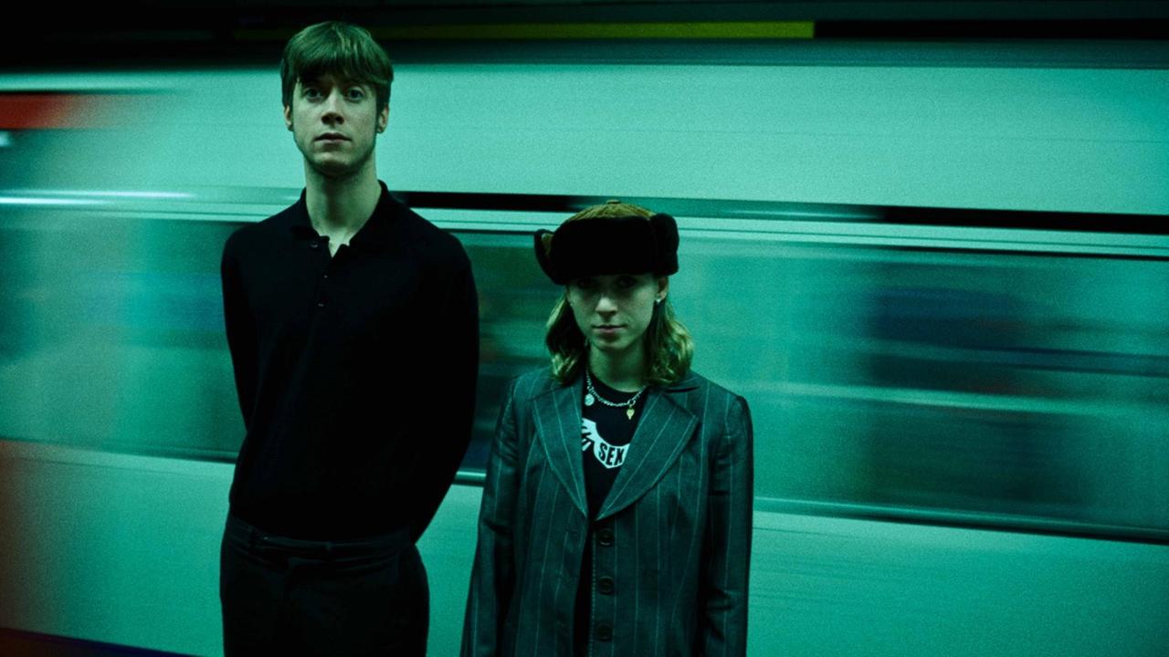 Ein Mann im dunklen Pulii und eine Frau mit Pelzmütze und Jacket stehen vor einem vorbeifahrendem Zug.