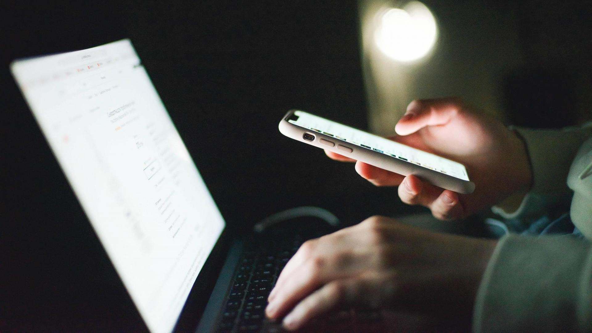 Eine Frau blickt auf einen Laptop und hält gleichzeitig ein eingeschaltetes Smartphone in der Hand.