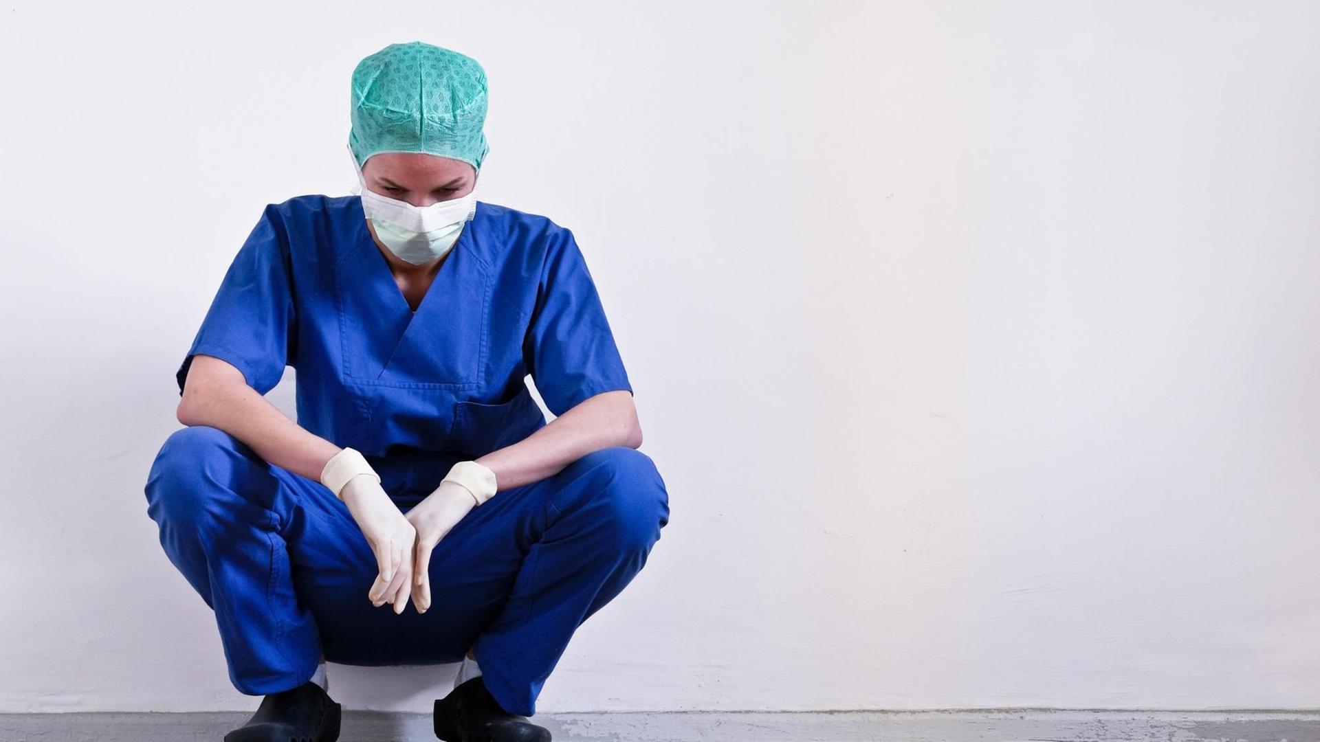 Eine erschöpfte Ärztin hockt noch in OP-Kleidung nach einer Operation am Boden. Eine erschöpfte Ärztin hockt noch in OP-Kleidung nach einer Operation am Boden.