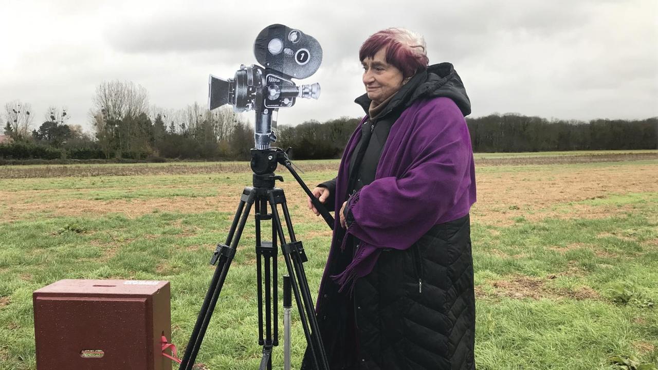 Agnes Varda steht auf einem Kamerawagen der auf Schienen in einer Landschaft steht. Agnes Varda steht auf einem Kamerawagen der auf Schienen in einer Landschaft steht.