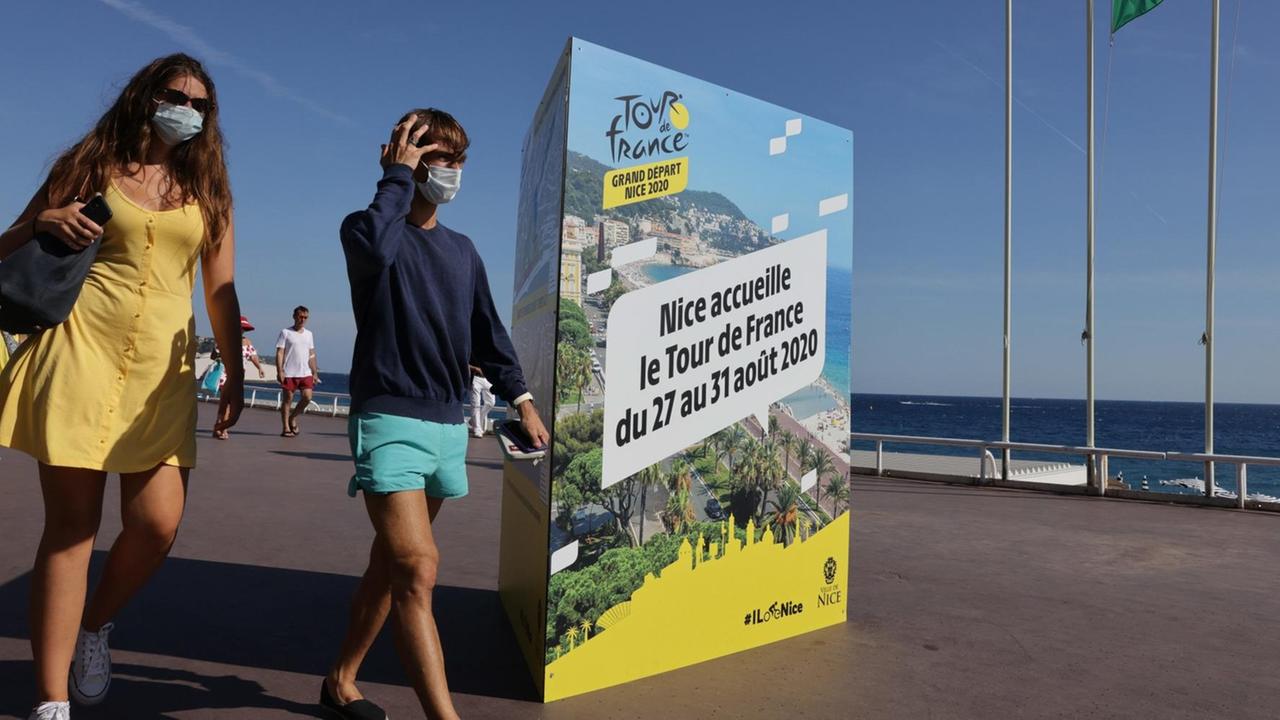 Zwei Passanten mit Mundschutz gehen auf der Promenade des Anglais in Nizza an einem Plakat vorbei, das die Tour de France 2020 ankündigt Zwei Passanten mit Mundschutz gehen auf der Promenade des Anglais in Nizza an einem Plakat vorbei, das die Tour de France 2020 ankündigt