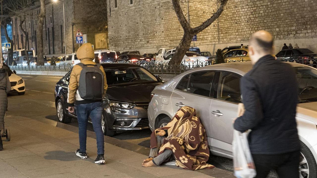 Ein barfüßiger Mann verhüllt von einer Decke sitzt auf dem Bürgersteig neben einem Auto und streckt die rechte Hand aus. Ein barfüßiger Mann verhüllt von einer Decke sitzt auf dem Bürgersteig neben einem Auto und streckt die rechte Hand aus.