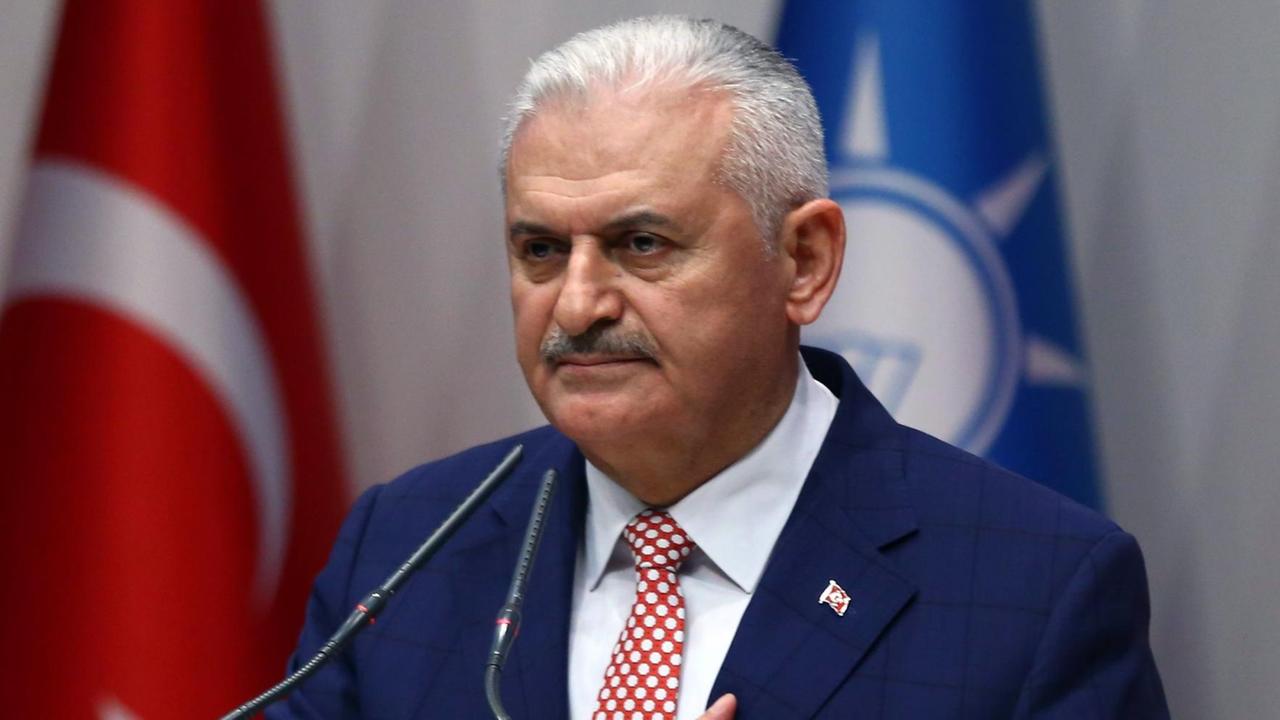 Binali steht vor zwei Mikrofonen. Hinter ihm sieht man die Fahnen der Türkei und der AKP. Binali steht vor zwei Mikrofonen. Hinter ihm sieht man die Fahnen der Türkei und der AKP.