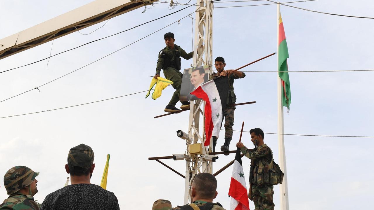 Syrische Regierungssoldaten klettern an der türkischen Grenze in der Stadt Kobane auf einen Strommast mit der Nationalflagge der syrischen Regierung und der gelben Fahne der Kurdischen Volksschutzeinheiten (YPG), zusammen mit einem Porträt des syrischen Präsidenten Bashar al-Assad. Im Hintergrund weht die gelb-rot-grünen kurdischen Rojava-Flagge auf einem Mast. Syrische Regierungssoldaten klettern an der türkischen Grenze in der Stadt Kobane auf einen Strommast mit der Nationalflagge der syrischen Regierung und der gelben Fahne der Kurdischen Volksschutzeinheiten (YPG), zusammen mit einem Porträt des syrischen Präsidenten Bashar al-Assad. Im Hintergrund weht die gelb-rot-grünen kurdischen Rojava-Flagge auf einem Mast.