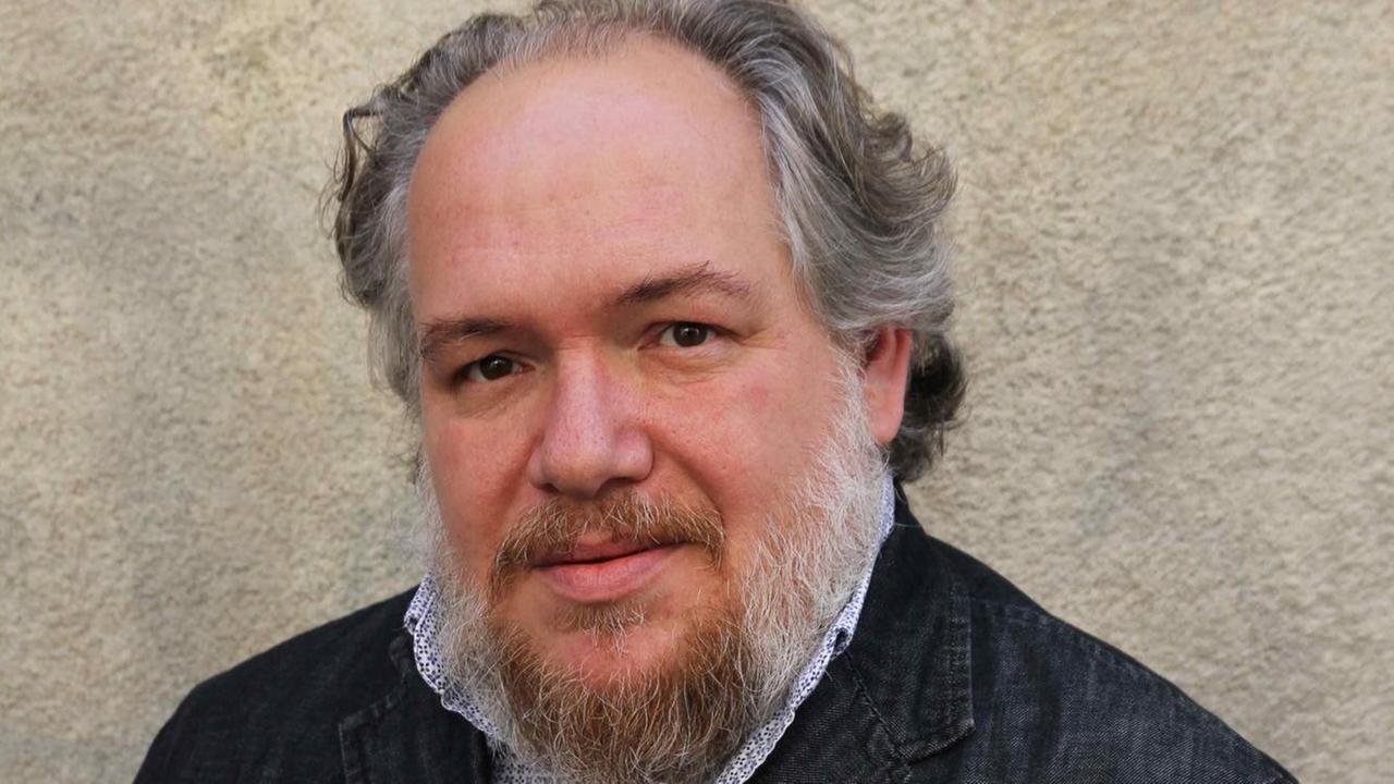 Mathias Enard hat grau-weiße Haare und trägt Vollbart. Er hat ein helles Hemd und eine dunkle Jacke an. Mathias Enard hat grau-weiße Haare und trägt Vollbart. Er hat ein helles Hemd und eine dunkle Jacke an.