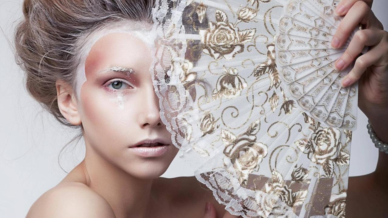 Eine Frau, weiß und gold geschminkt, hält einen Fächer, der ihr Gesicht teilweise verdeckt. Eine Frau, weiß und gold geschminkt, hält einen Fächer, der ihr Gesicht teilweise verdeckt.