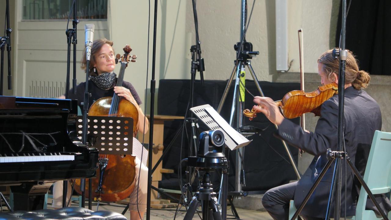 Frau mit Cello und Mann mit Geige stimmen ihre Instrumente zwischen Mikrofonständern