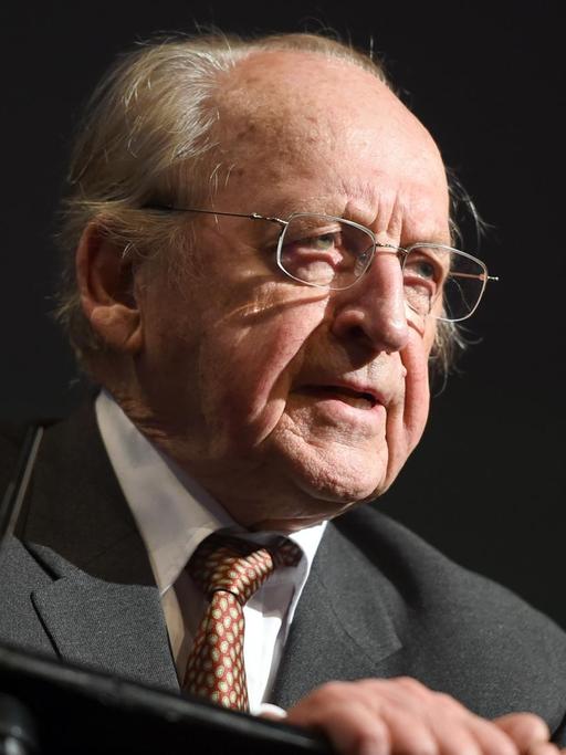 Günther Rühle steht im Dunklen an einem Rednerpult und spricht, nur auf ihm liegt Scheinweiferlicht.