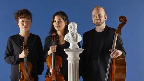 Die Musiker stehen mit ihren Instrumenten vor einer blauen Wand. Vor ihnen auf einem weißen Sockel eine kleine Statue von Beethoven.