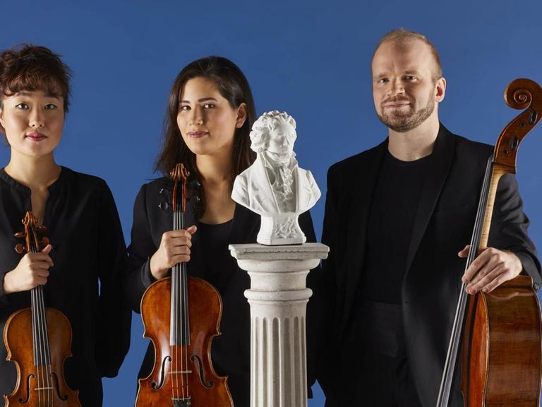 Die Musiker stehen mit ihren Instrumenten vor einer blauen Wand. Vor ihnen auf einem weißen Sockel eine kleine Statue von Beethoven.