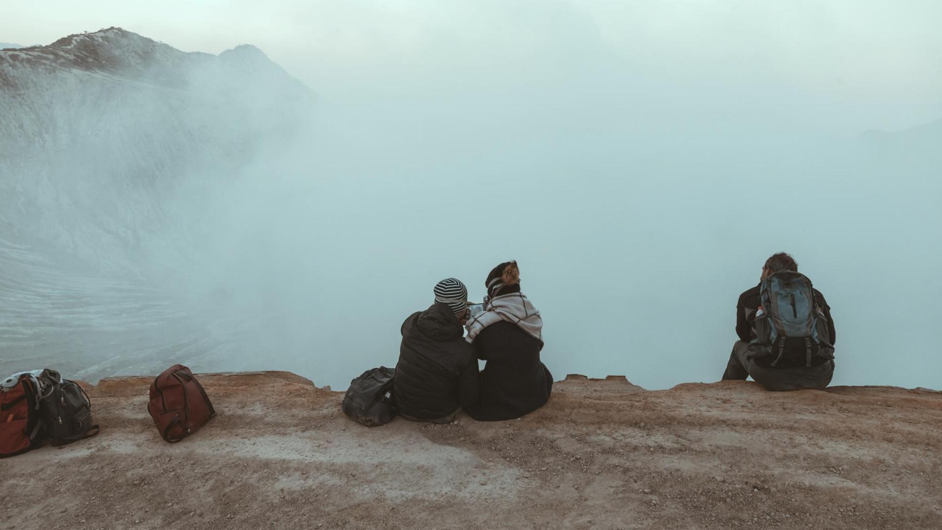Drei junge Menschen sitzen mit Rucksäcken auf einem Berg und schauen in die Wolken.