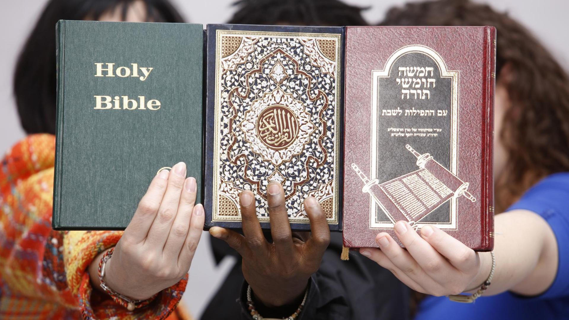 Drei Personen halten eine Bibel, einen Koran und ein Buch mit jüdischen Schriften.