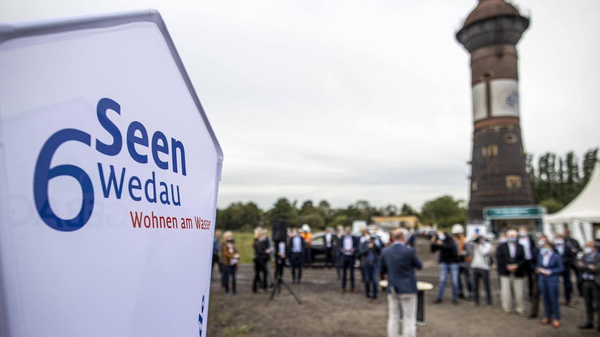 Im Vordergrund ein Schild, das auf das Projekt 6 Seen Wedau hinweist, im Hintergrund sind versammelte Menschen und der Wasserturm des alten Rangierbahnhofs zu sehen. Im Vordergrund ein Schild, das auf das Projekt 6 Seen Wedau hinweist, im Hintergrund sind versammelte Menschen und der Wasserturm des alten Rangierbahnhofs zu sehen.