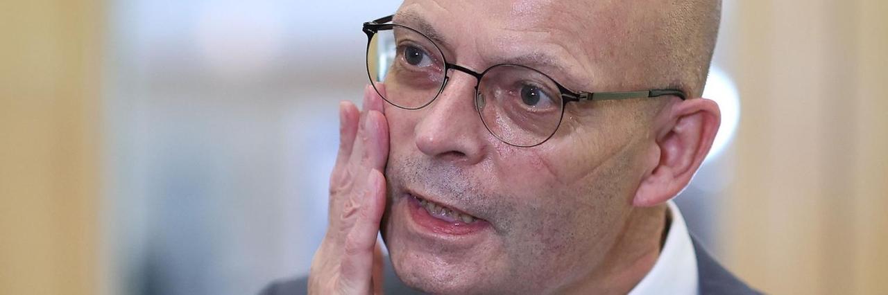 Wiegand mit Brille im schwarzen Anzug sagt etwas und hält dabei seine rechte Hand an die Backe. Wiegand mit Brille im schwarzen Anzug sagt etwas und hält dabei seine rechte Hand an die Backe.
