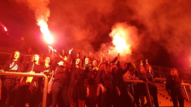 Weibliche Ultrafans aus ganz Europa zur Eröffnung der Ausstellung "Fan.Tastic Females". Eine Gruppe Frauen steht in einem Fußballstadion, das durch Feuer beleuchtet wird.
