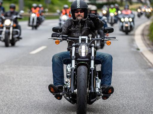 Auf dem Foto fahren zahlreiche Harley Davidson-Fahrer auf den Fotografen zu. Im Vordergrund ist ein Fahrer mit einem gezwirbelten grauen Backenbart im Gesicht.