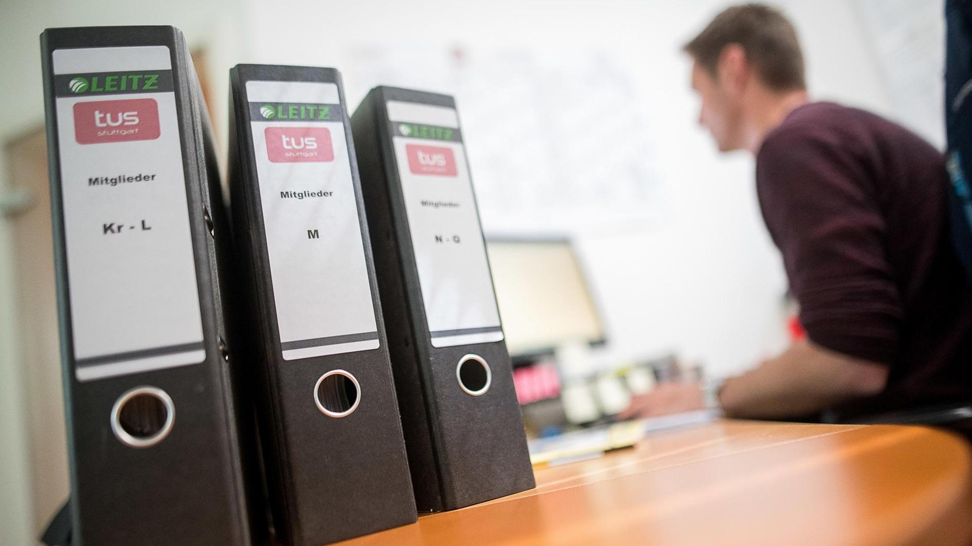 22.05.2018, Baden-Württemberg, Stuttgart: Aktenordner mit Mitgliedsanträgen des Sportvereins TUS Stuttgart stehen auf einem Tisch, während im Hintergrund ein Mann an einem Computer arbeitet. Am 25. Mai tritt die neue EU-Datenschutzregel in Kraft. Vereine und Unternehmen klagen über einen großen Aufwand. Datenschützer loben den besseren Schutz von personenbezogenen Daten der Bürger. (zu dpa "Neue Datenschutzregeln treiben Vereine und Unternehmen um" vom 23.05.2018) Foto: Sebastian Gollnow/dpa | Verwendung weltweit 22.05.2018, Baden-Württemberg, Stuttgart: Aktenordner mit Mitgliedsanträgen des Sportvereins TUS Stuttgart stehen auf einem Tisch, während im Hintergrund ein Mann an einem Computer arbeitet. Am 25. Mai tritt die neue EU-Datenschutzregel in Kraft. Vereine und Unternehmen klagen über einen großen Aufwand. Datenschützer loben den besseren Schutz von personenbezogenen Daten der Bürger. (zu dpa "Neue Datenschutzregeln treiben Vereine und Unternehmen um" vom 23.05.2018) Foto: Sebastian Gollnow/dpa | Verwendung weltweit