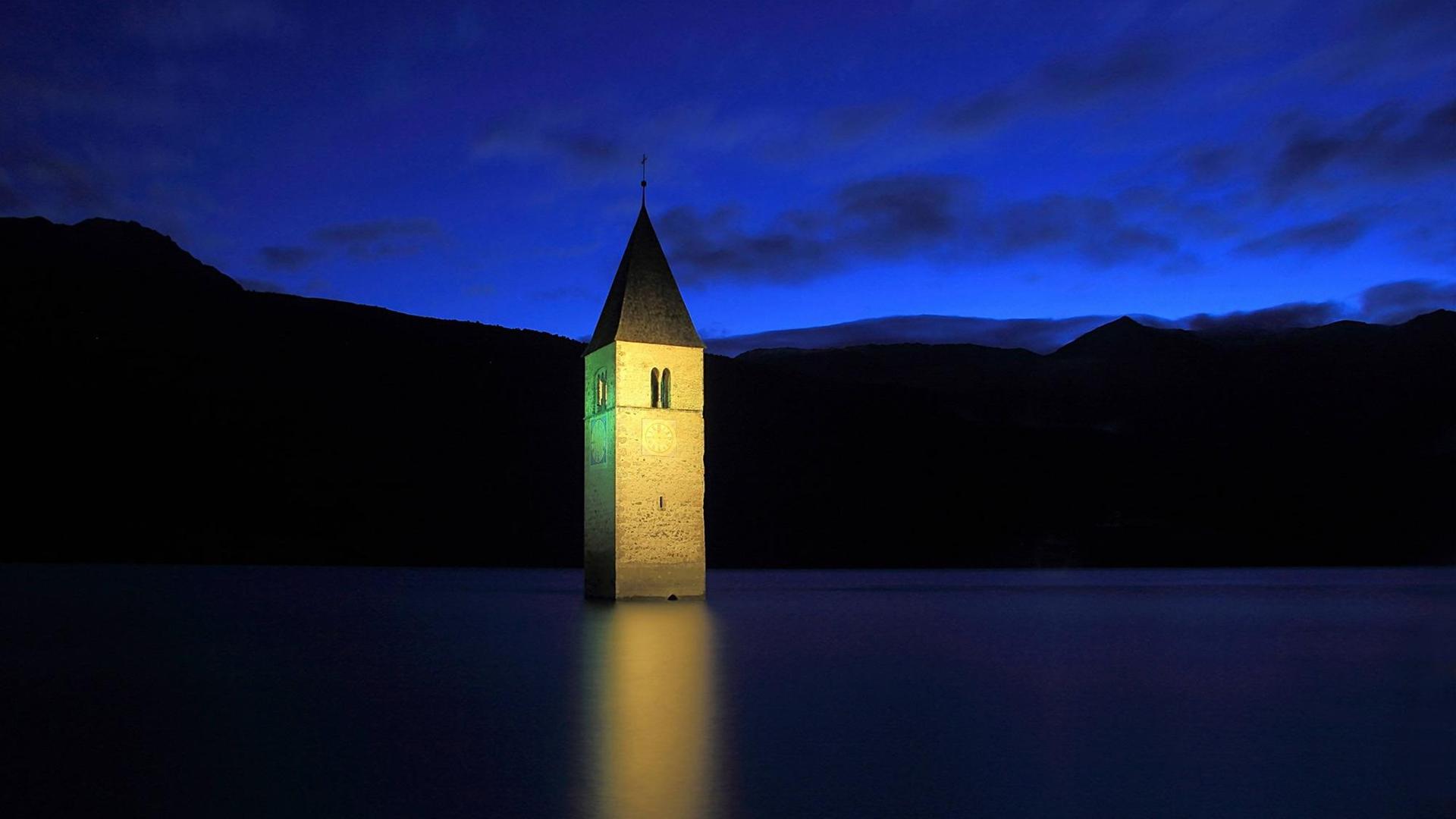 Der Kirchturm des Dorfes Graun ragt im Nachtlicht aus dem Wasser eines Stausees. Das Dorf Graun wurde nach dem Zweiten Weltkrieg von der italienischen Zentralregierung geflutet. Der Kirchturm des Dorfes Graun ragt im Nachtlicht aus dem Wasser eines Stausees. Das Dorf Graun wurde nach dem Zweiten Weltkrieg von der italienischen Zentralregierung geflutet.