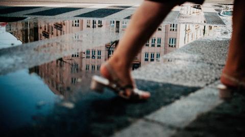 Hinter den Beinen einer vorbeiflanierenden Frau spiegeln sich in einer Pfütze die Fassaden der umliegenden Gebäude.