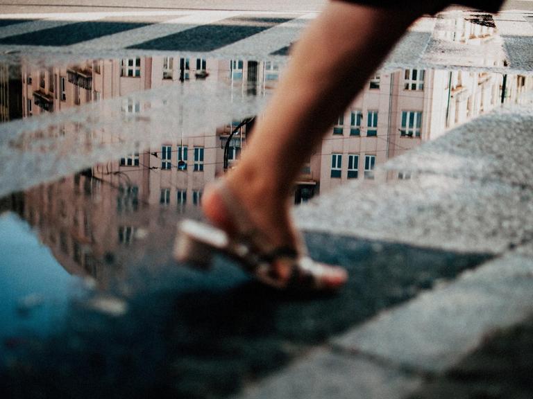 Hinter den Beinen einer vorbeiflanierenden Frau spiegeln sich in einer Pfütze die Fassaden der umliegenden Gebäude.