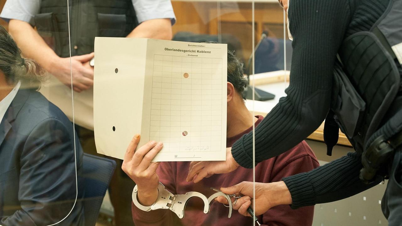 Ein Mann im Gericht hält sich eine Akte vors Gesicht, während ihm ein Beamter die Handschellen abnimmt. Ein Mann im Gericht hält sich eine Akte vors Gesicht, während ihm ein Beamter die Handschellen abnimmt.