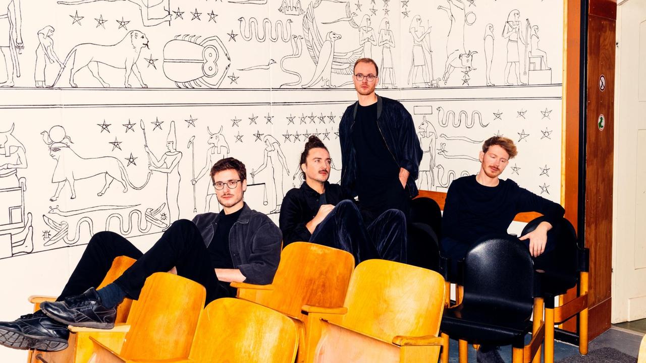 Vier junge Musiker lümmeln in hölzernen Kinostühlen vor weißer Wand mit ägyptischen Motiven Vier junge Musiker lümmeln in hölzernen Kinostühlen vor weißer Wand mit ägyptischen Motiven