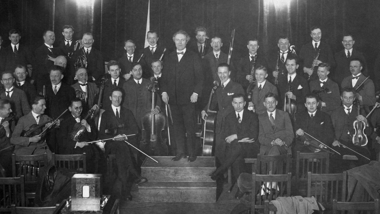 Auf einer historischen Fotografie steht ein Dirigent von Musikern umgeben auf einem erhöhten Pult. 