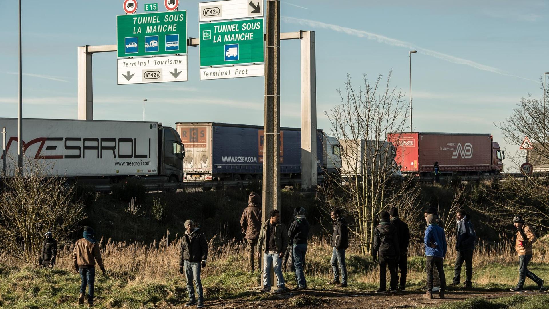 Flüchtlinge stehen am Rande einer Autobahn, auf der Lkws fahren. Über der Straße weist ein Schild Richtung Eurotunnel. Flüchtlinge stehen am Rande einer Autobahn, auf der Lkws fahren. Über der Straße weist ein Schild Richtung Eurotunnel.