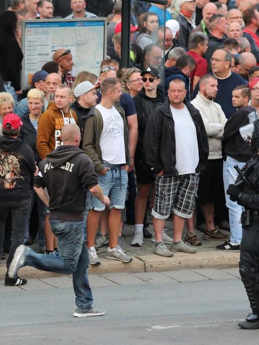 Demonstranten stehen am 27.08.2018 am Karl-Marx-Monument in Chemnitz um eine Bushaltestelle herum, im Vordergrund haben sich vier Polizisten positioniert.