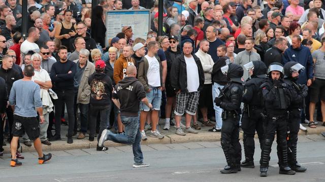 Demonstranten stehen am 27.08.2018 am Karl-Marx-Monument in Chemnitz um eine Bushaltestelle herum, im Vordergrund haben sich vier Polizisten positioniert.