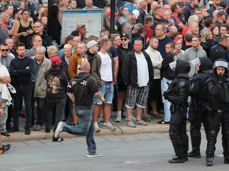 Demonstranten stehen am 27.08.2018 am Karl-Marx-Monument in Chemnitz um eine Bushaltestelle herum, im Vordergrund haben sich vier Polizisten positioniert.