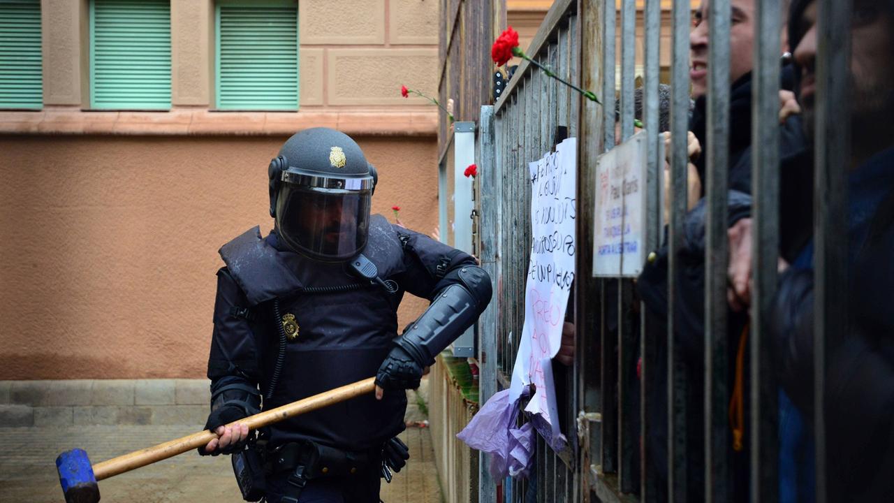 Spanische Polizisten versuchen in die Pau Claris Schule in Barcelona einzudringen, in der über das katalonische Referendum abgestimmt wird. Man sieht einen Polizisten mit schussicherer Weste und mit geschlossenem Helm, der mit einem riesigen Hammer vor dem vergitterten Eingang der Schule steht und offenbar zum Schlag ausholt. Spanische Polizisten versuchen in die Pau Claris Schule in Barcelona einzudringen, in der über das katalonische Referendum abgestimmt wird. Man sieht einen Polizisten mit schussicherer Weste und mit geschlossenem Helm, der mit einem riesigen Hammer vor dem vergitterten Eingang der Schule steht und offenbar zum Schlag ausholt.