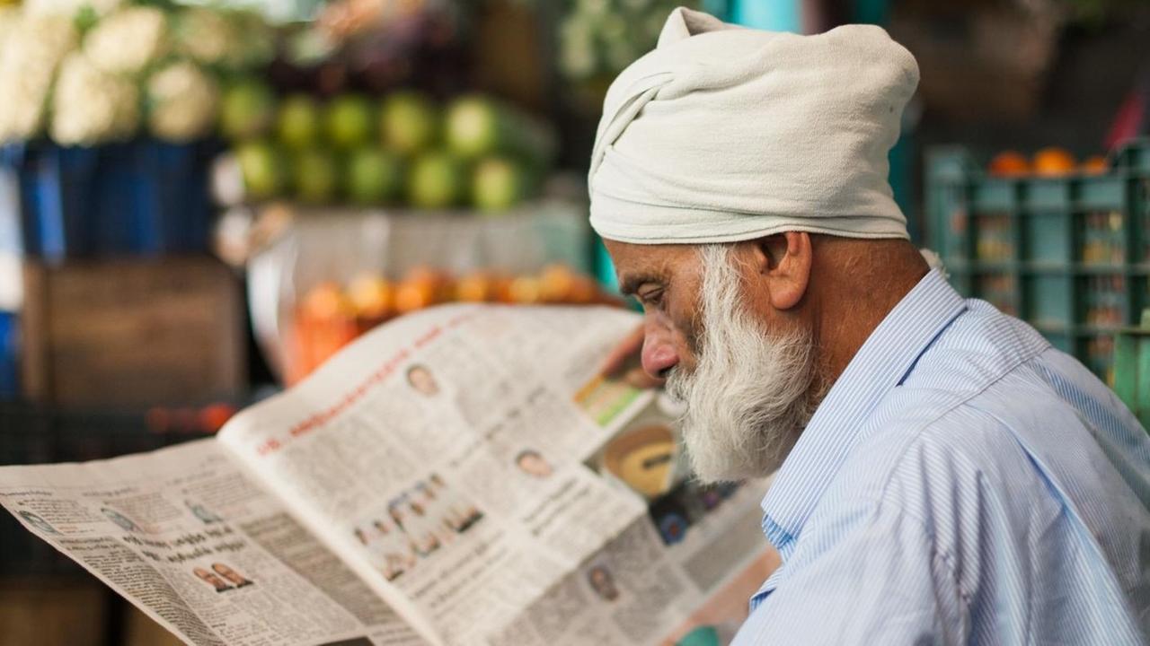 Der alte Mann mit Zeitung trägt einen weißen Bart und einen weißen Turban. Er sitzt vor einem Marktstand mit Obst. Der alte Mann mit Zeitung trägt einen weißen Bart und einen weißen Turban. Er sitzt vor einem Marktstand mit Obst.