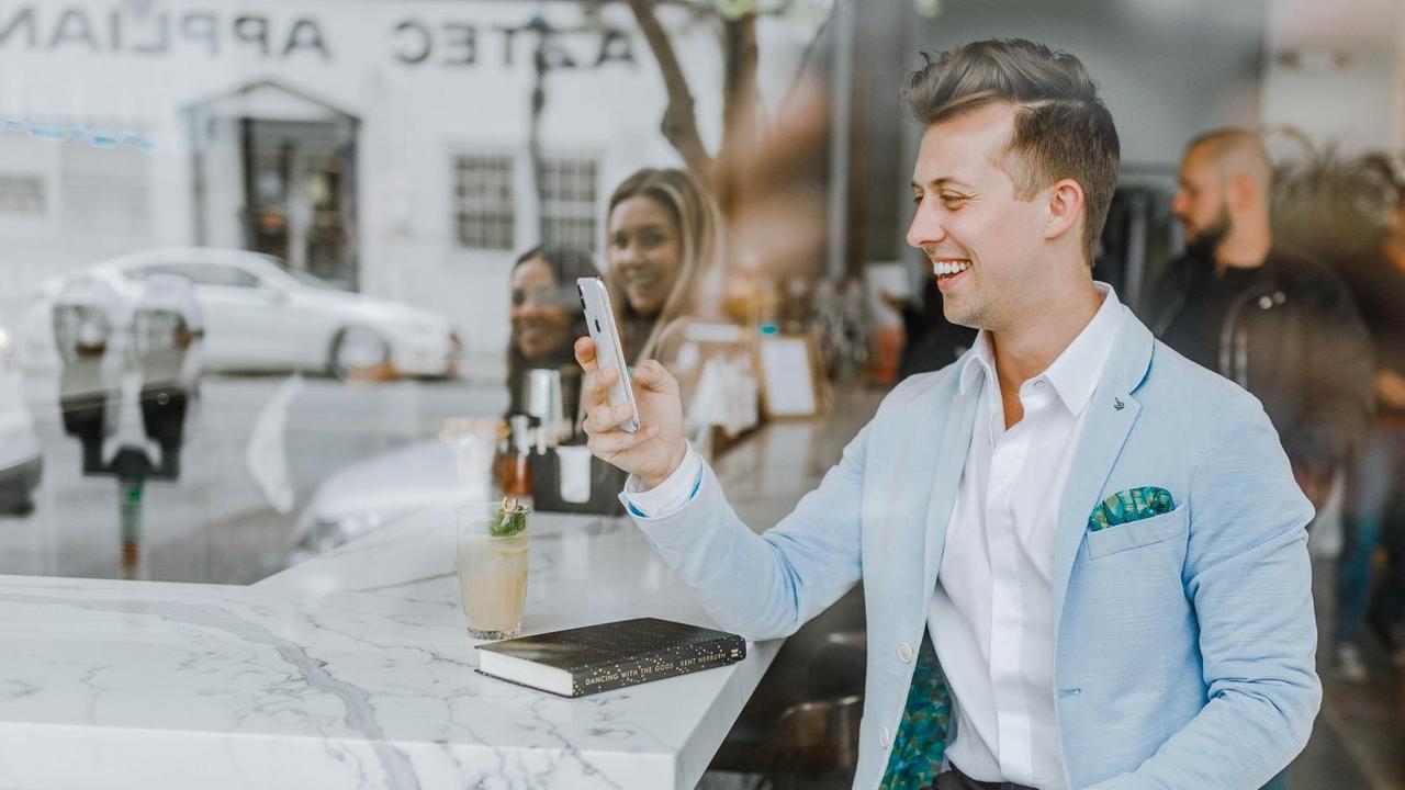 Ein junger, gepflegter Mann in gutem Anzug sitzt am Tresen eines schicken Cafés und lacht beim Blick auf sein Handy. 